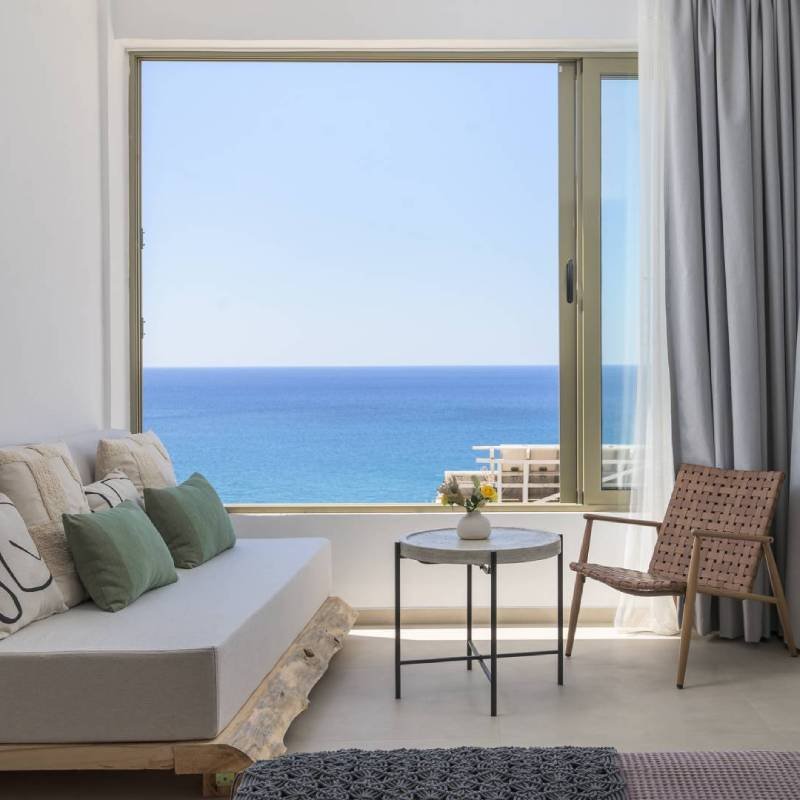 Interior of a bright, modern room featuring a comfortable sofa and chair facing a large window with unobstructed sea view. Showcases the relaxing accommodation services at Panorama Myrtos, Crete.