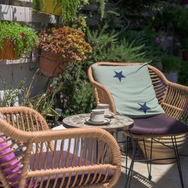 Close-up of cozy wicker chairs with patterned cushions in a lush, sunny garden setting, symbolizing the warm welcome and tranquil atmosphere at Panorama Myrtos, Crete.