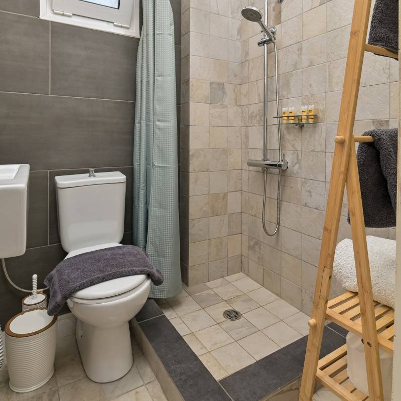 Spotless Modern Bathroom & Premium Toiletries - Panorama Myrtos Modern, clean bathroom with walk-in shower, tiled walls, fresh towels on a wooden ladder shelf, and premium toiletries. Highlights hygiene and comfort at Panorama Myrtos.