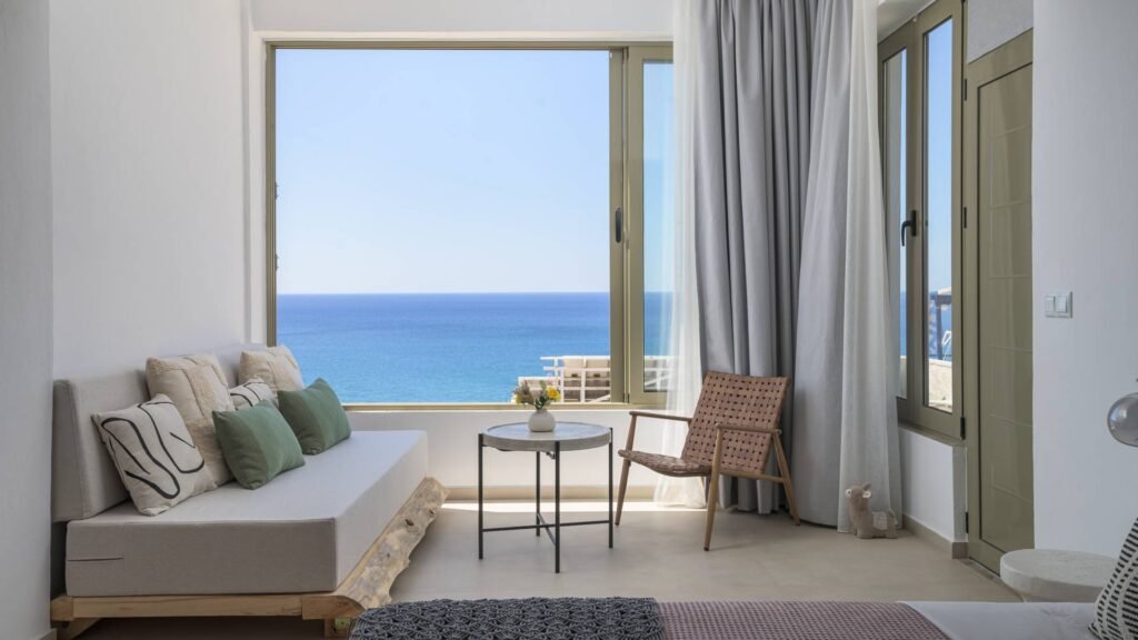 Luxury Sea View Apartments in Myrtos, Crete - Panorama Modern, luxury apartment interior with a cozy sofa and chair, featuring a large window with an **unobstructed, panoramic sea view** of the Mediterranean Sea in Myrtos, Crete