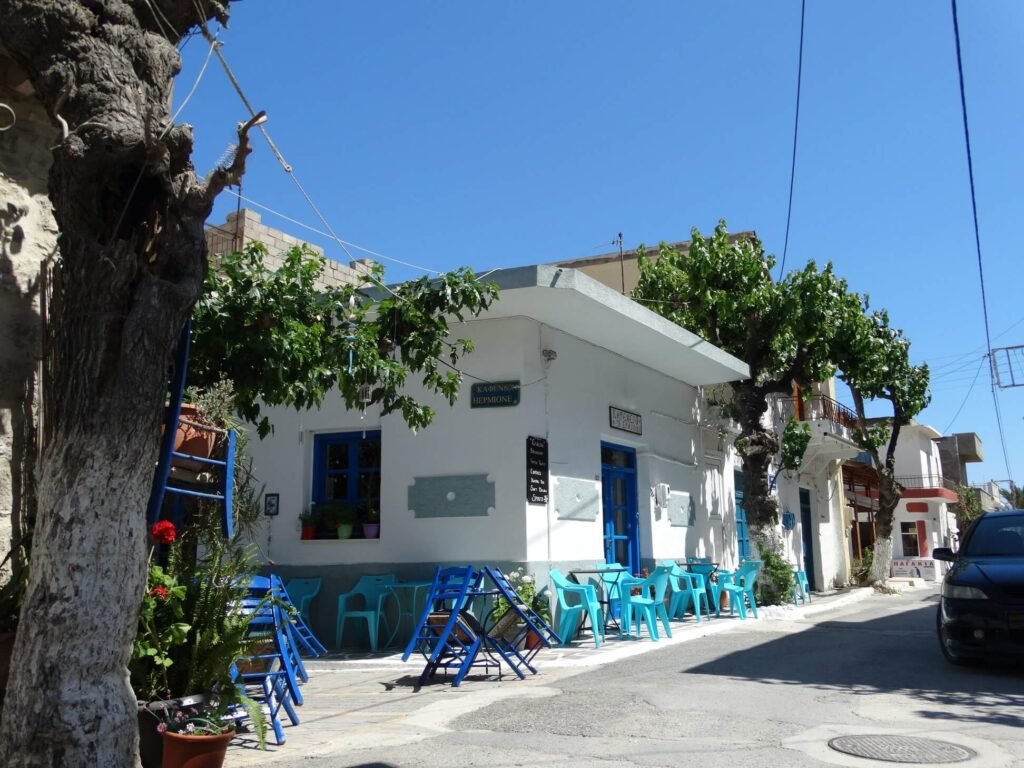 Traditional Cretan Kafeneio (coffee shop) in Myrtos village with white walls and bright blue chairs under green trees. Highlights local charm and dining options near Panorama Myrtos.