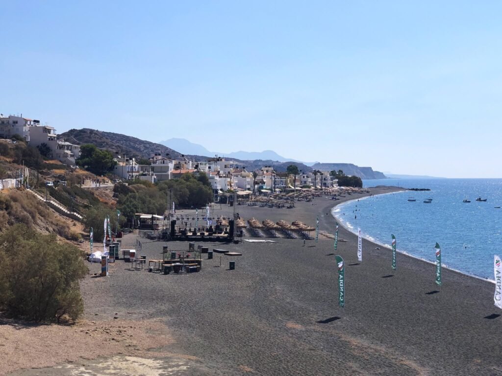 Wide view of the black sand Myrtos Beach in Crete, showing the seafront village, sunbeds, and clear blue water under a sunny sky. Highlights the prime location of Panorama Myrtos.